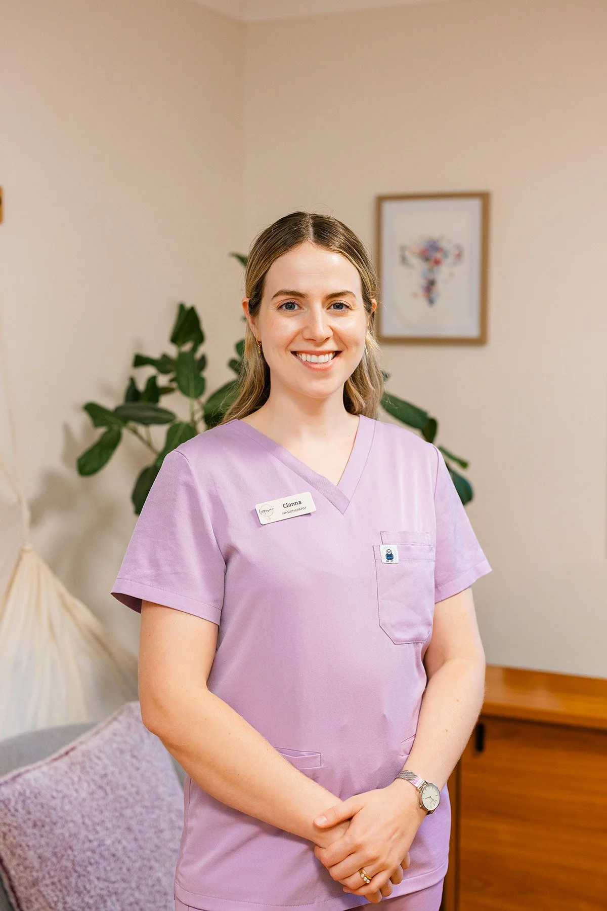 A woman in a lavender medical uniform smiling in an indoor setting with a green plant and framed artwork in the background.