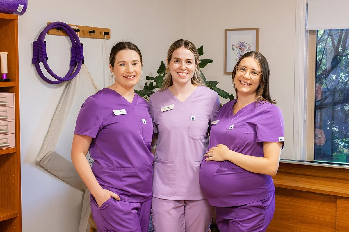 Three women in purple scrubs smiling in a medical office with plants and artwork in the background.