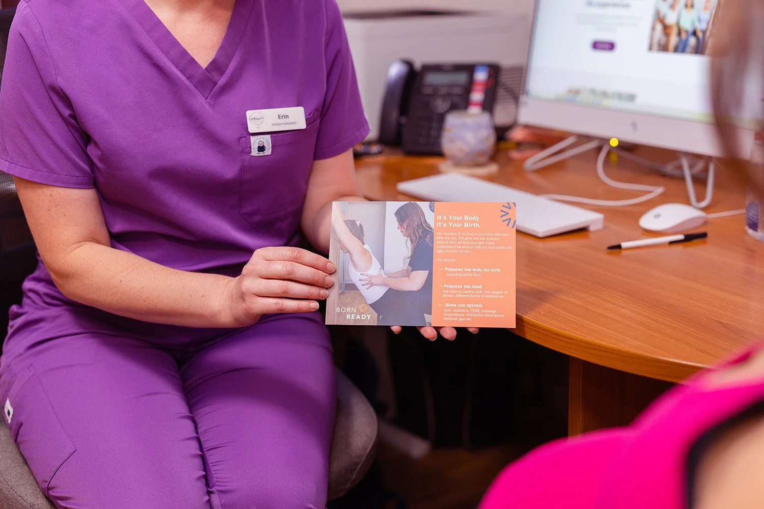 A woman in purple scrubs sitting at a desk holding an informational pamphlet about childbirth. The desk has a computer, keyboard, mouse, and some office supplies.