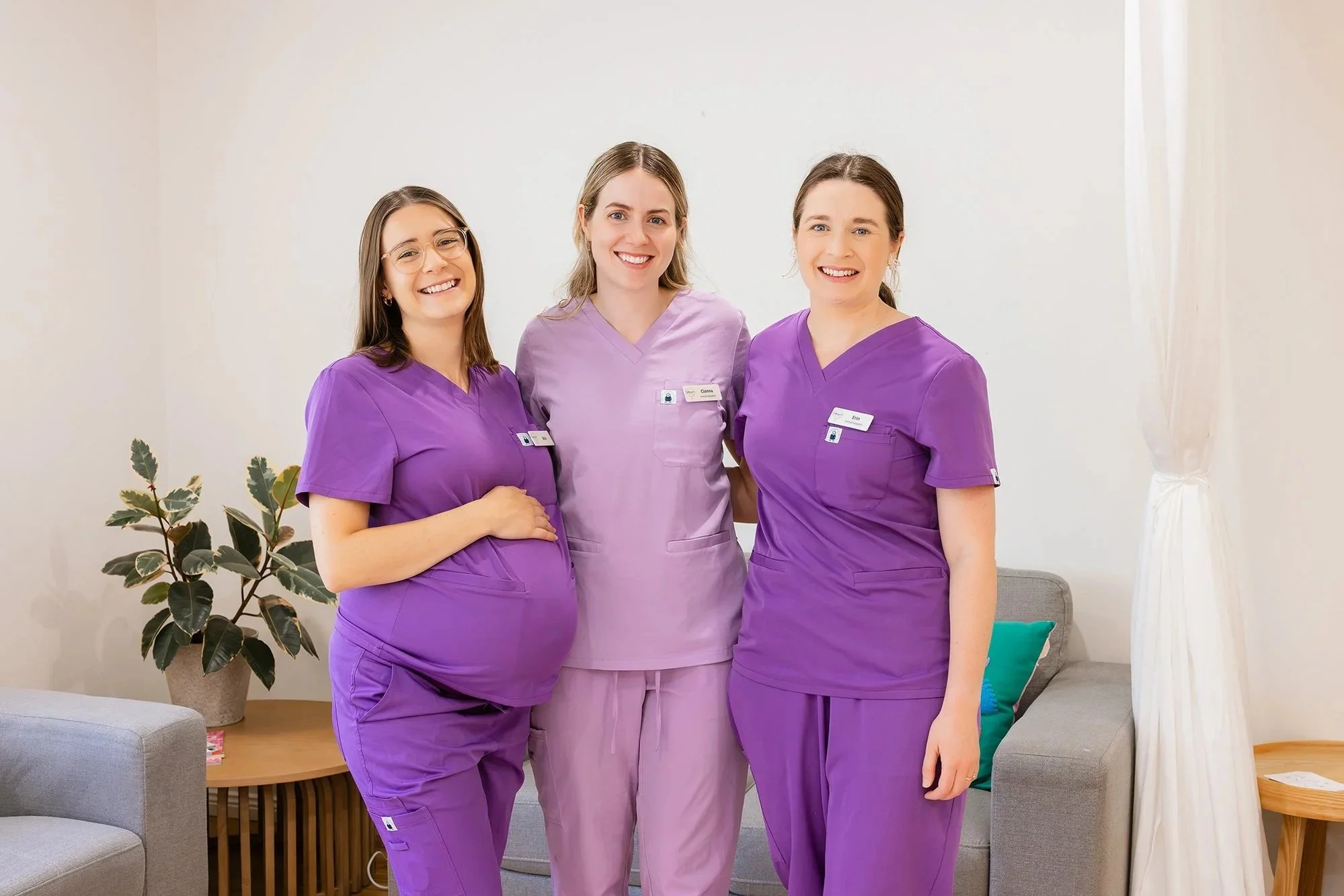 Three women wearing purple scrubs standing together in a room with a beige wall, a gray sofa, and a potted plant.
