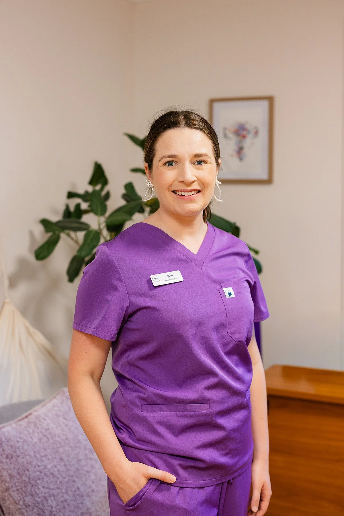 A smiling woman in purple scrubs with a name tag that says Erin, standing in a room with a large green plant, a framed colorful artwork on the wall, and a wooden table in the background.