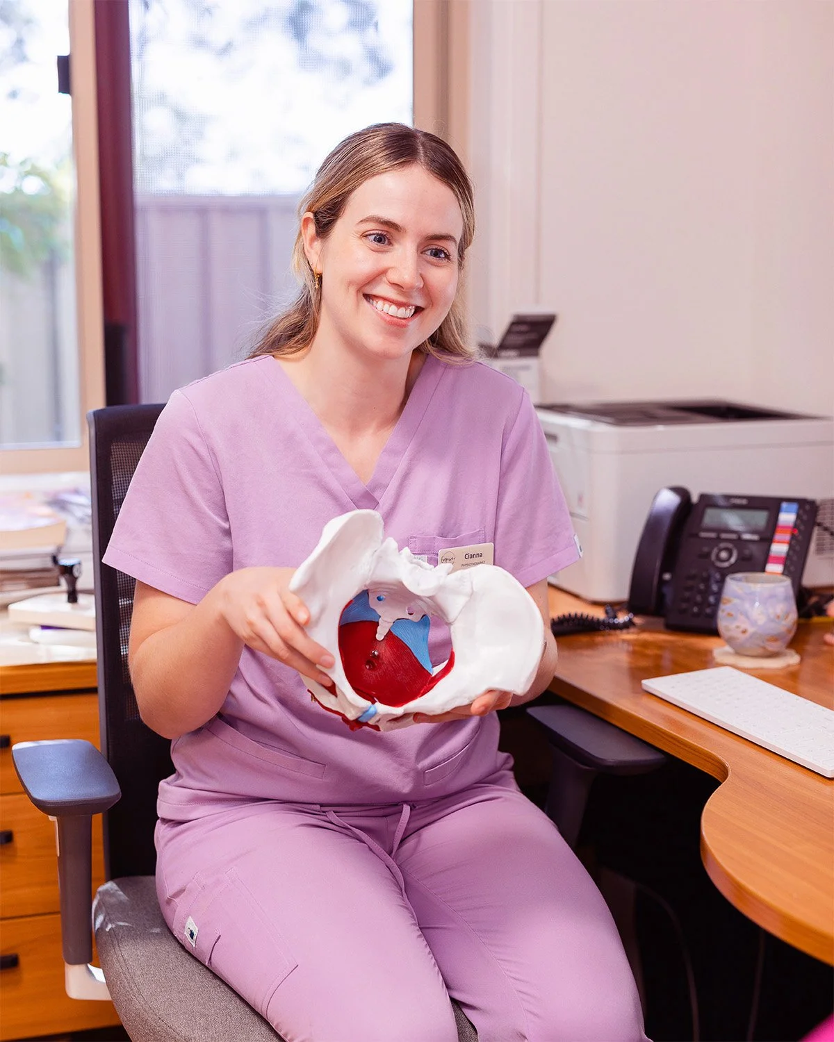 A smiling woman in pink scrubs holding a medical model of a pelvis with internal organs inside, sitting at a desk in an office.
