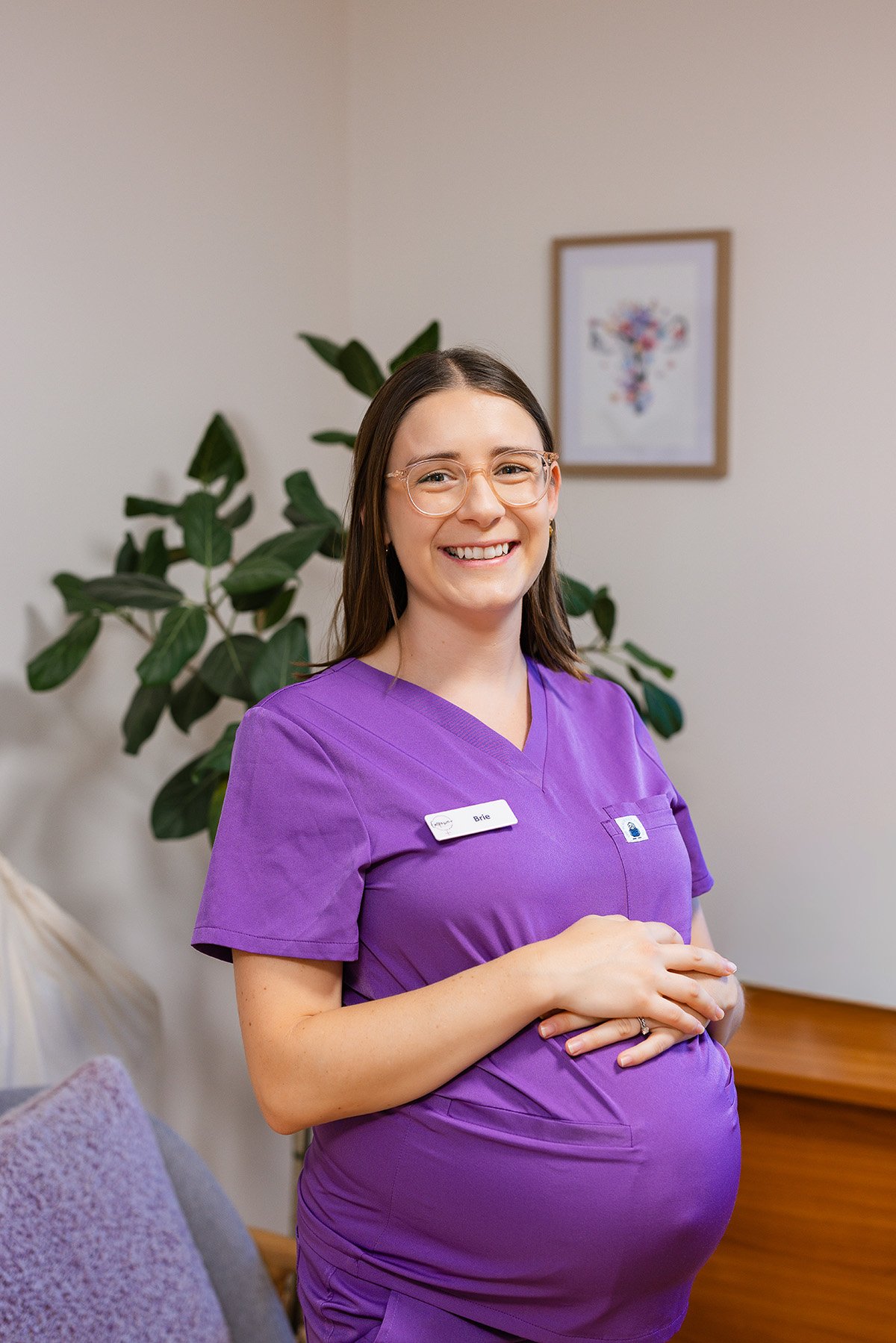 A smiling pregnant woman with glasses wearing purple medical scrubs and a name badge standing indoors with a houseplant and framed art on the wall behind her.