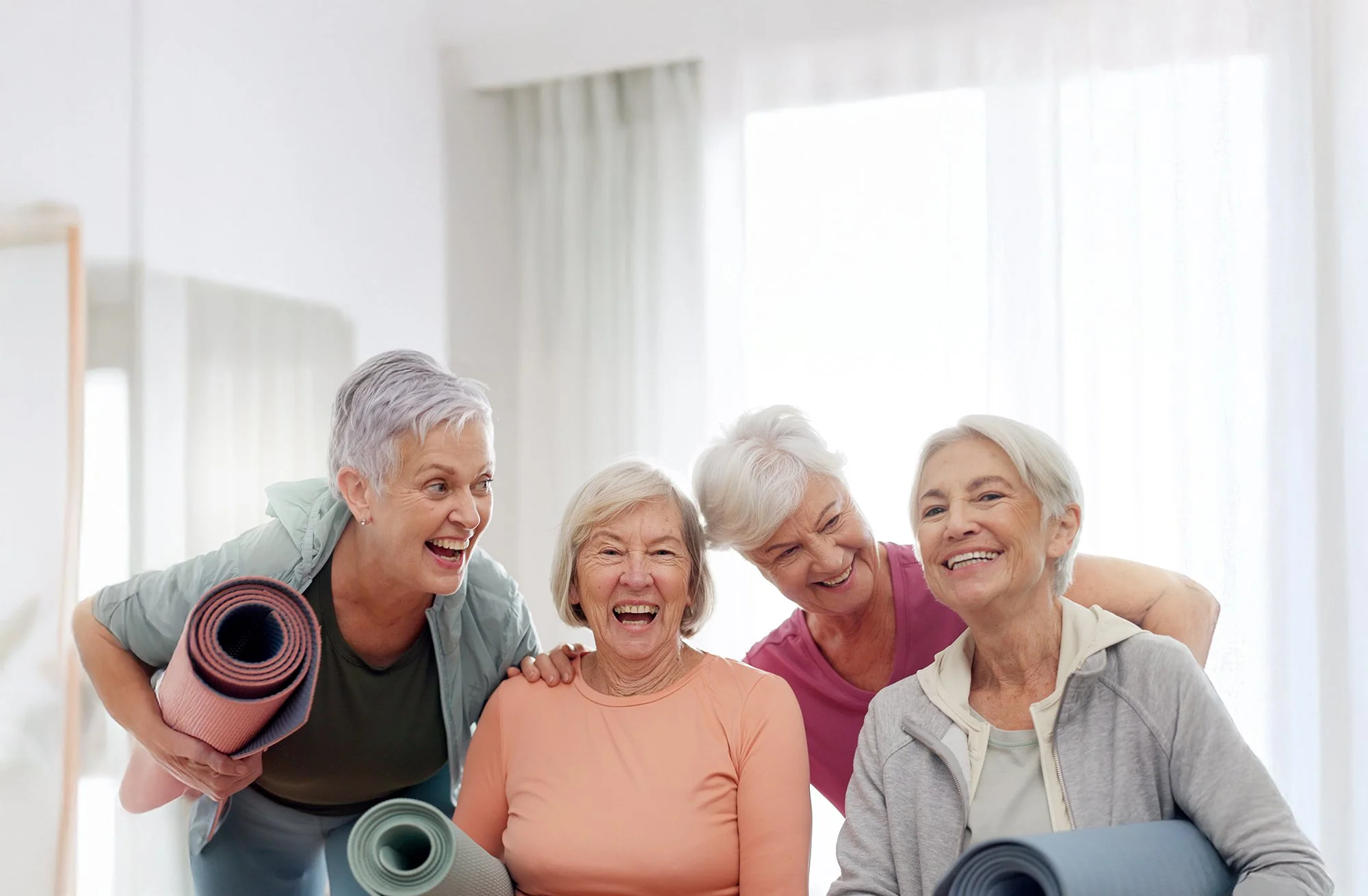 A group of five senior women smiling and laughing together, dressed in casual workout clothes, with some holding yoga mats, in a bright room with natural light.