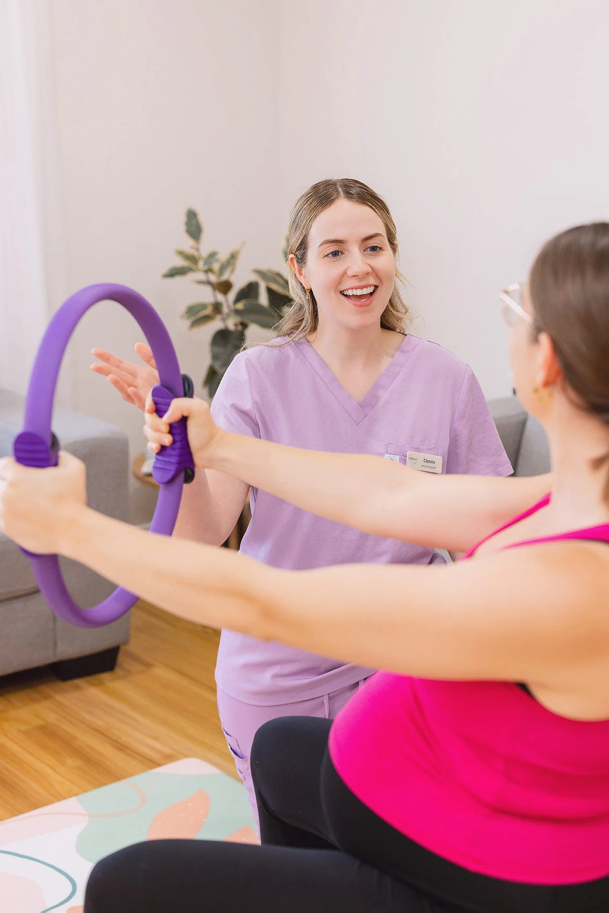 A healthcare worker in purple scrubs smiling as she assists a woman in a pink top with an exercise using a purple resistance ring.
