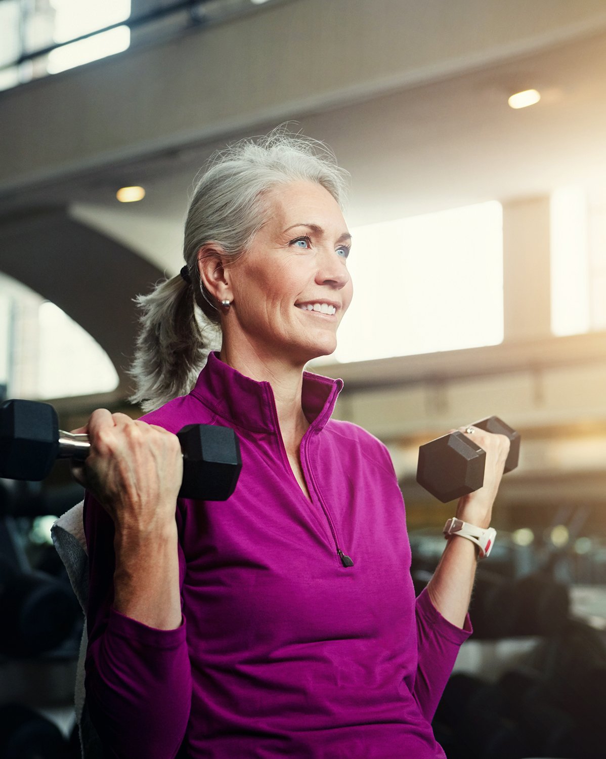 An elderly woman with gray hair pulled back, wearing a purple workout top, lifting dumbbells in a gym.