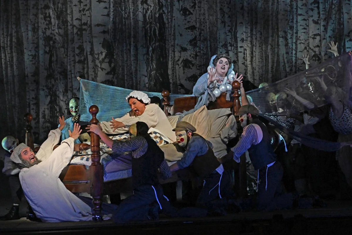 A theatrical performance with actors in clown makeup dressed as demons and people in old-fashioned clothing. Two women are in bed, one reaching out and the other appearing frightened while surrounded by actors in dark clothing.