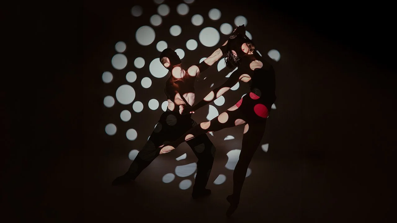 Two dancers silhouette against a dark background with white circular light projection, holding hands in a dance pose.