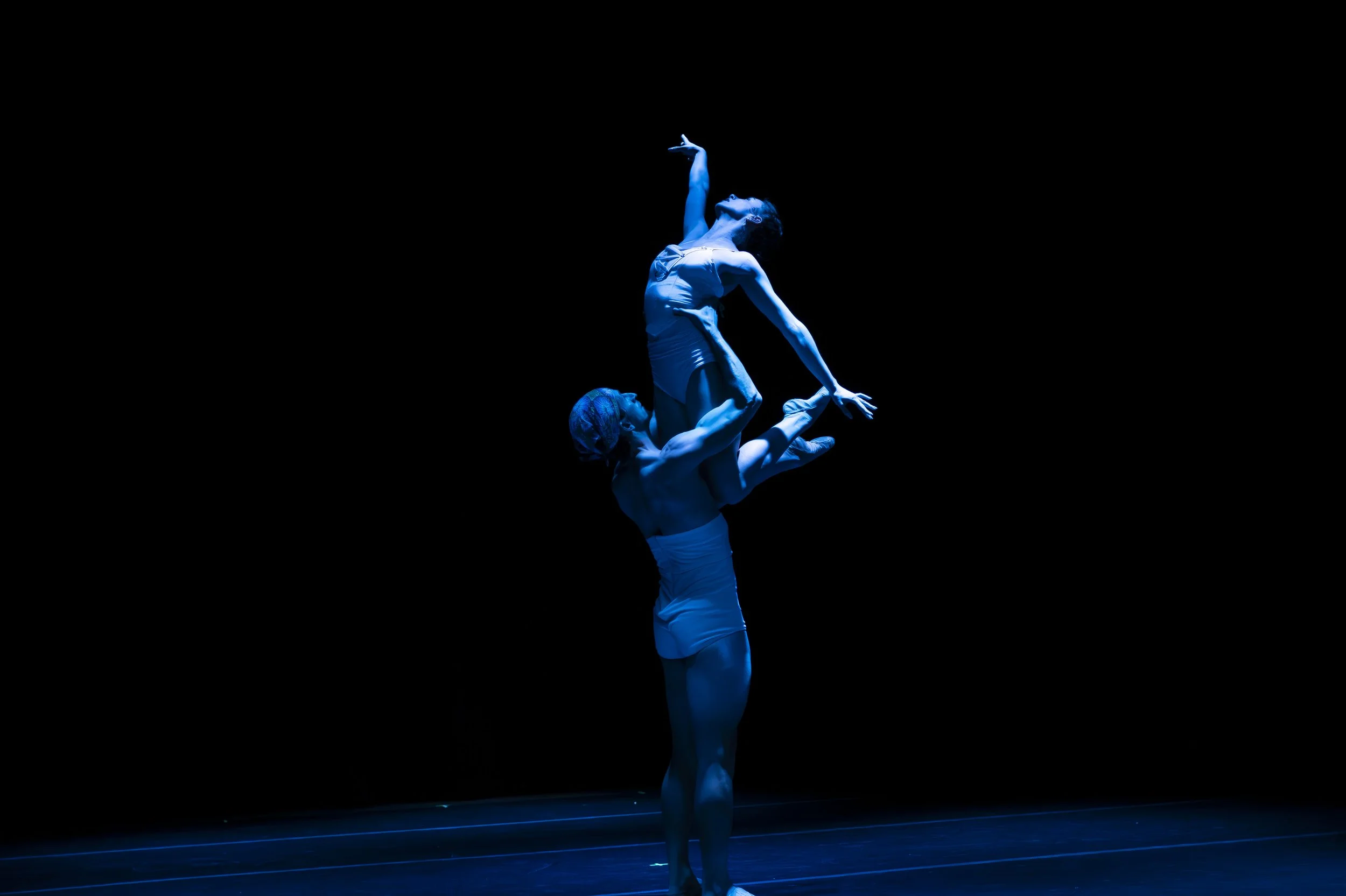 Two ballet dancers performing on stage with black background, illuminated in blue light, one standing and lifting the other in an intricate pose.