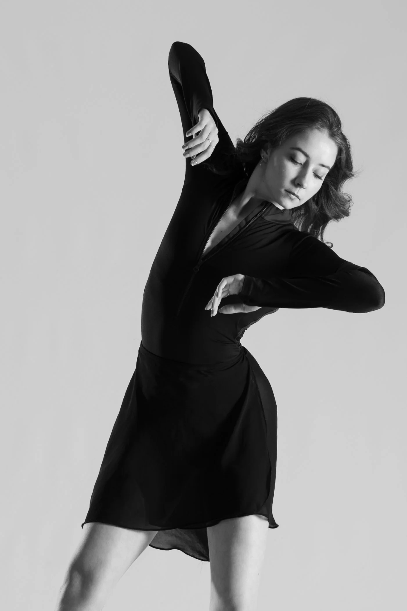 A woman with wavy hair dancing, wearing a black long-sleeve top and a black skirt, with her eyes closed and arms raised.