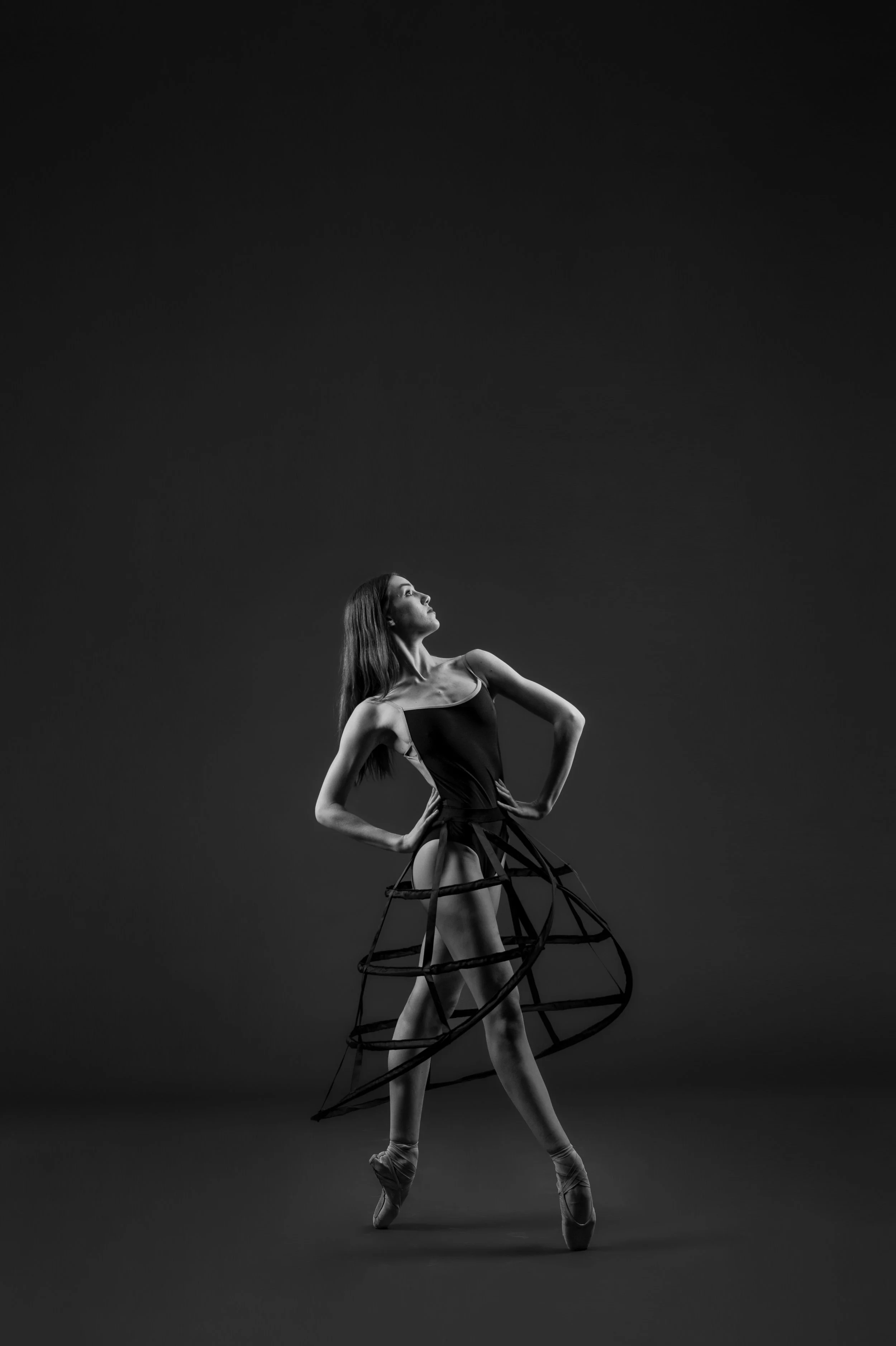 Black and white photo of a ballet dancer standing en pointe with her hands on her hips, wearing a dress with a cage-like skirt