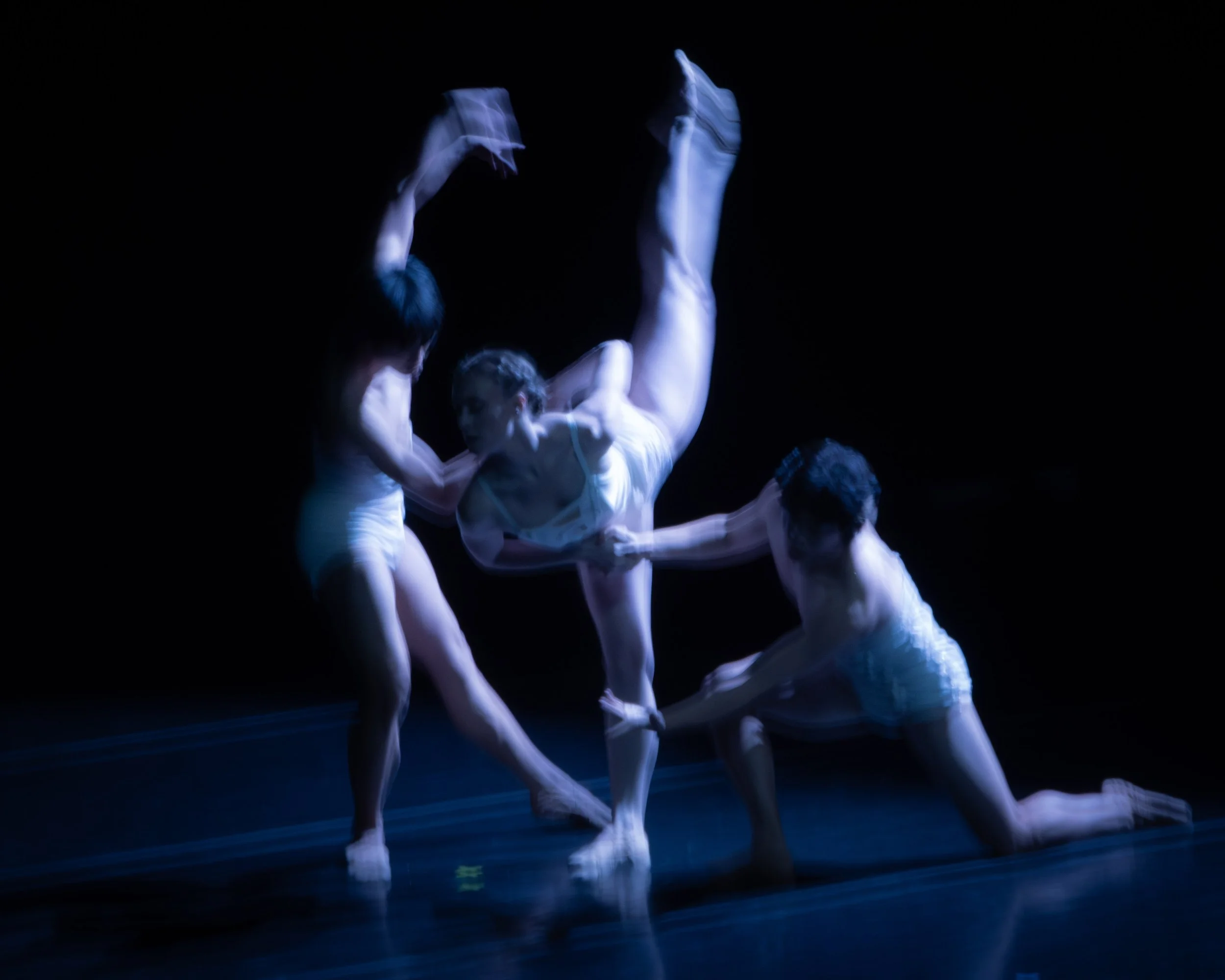 Three dancers performing a contemporary dance routine on stage, with one dancer in a handstand position and the other two supporting and holding her, dressed in white costumes, against a dark background.