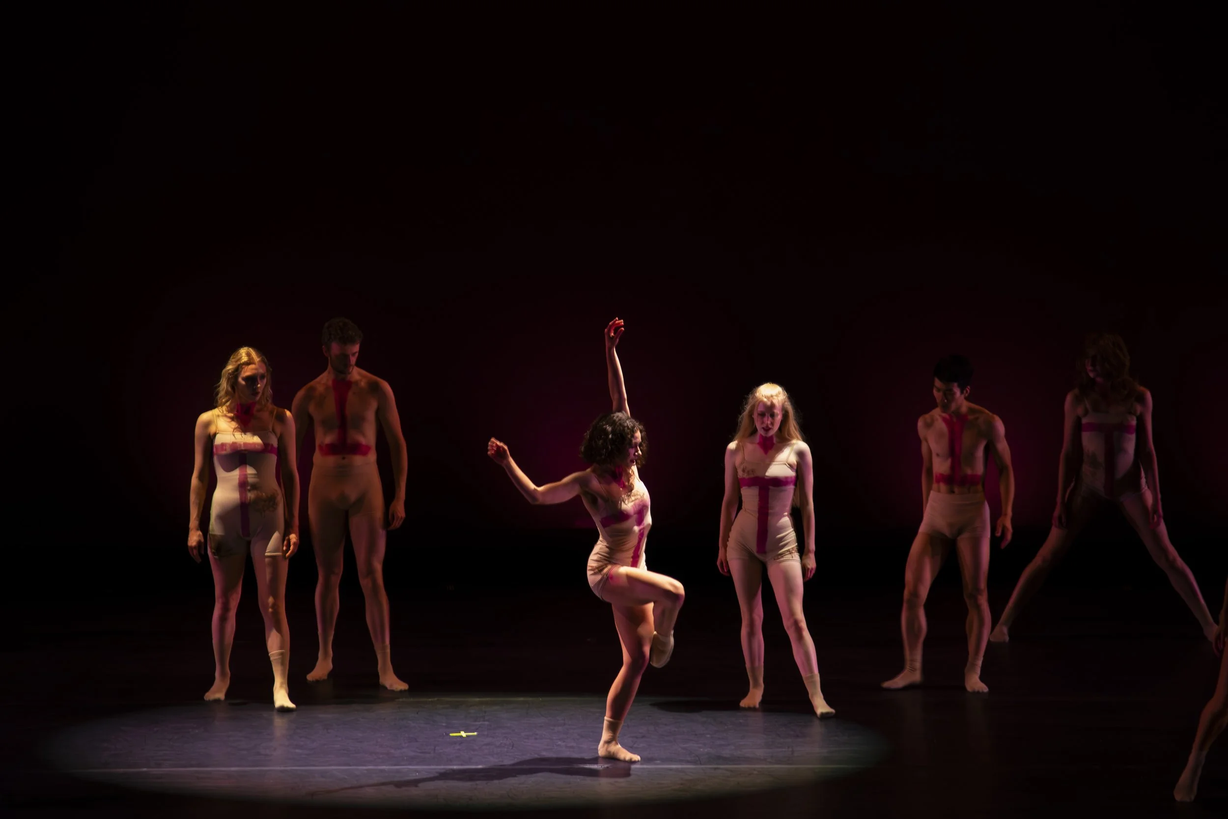 A group of six dancers on stage, five standing and one in mid-dance pose, wearing nude-colored costumes with red markings, illuminated by a spotlight on a dark stage.