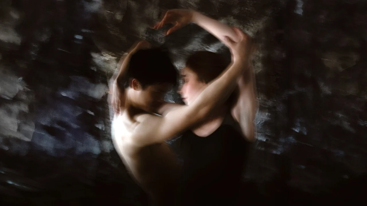 Two people, one shirtless and the other wearing a black shirt, are closely facing each other with their arms raised and touching above their heads, in a dark, rocky setting.