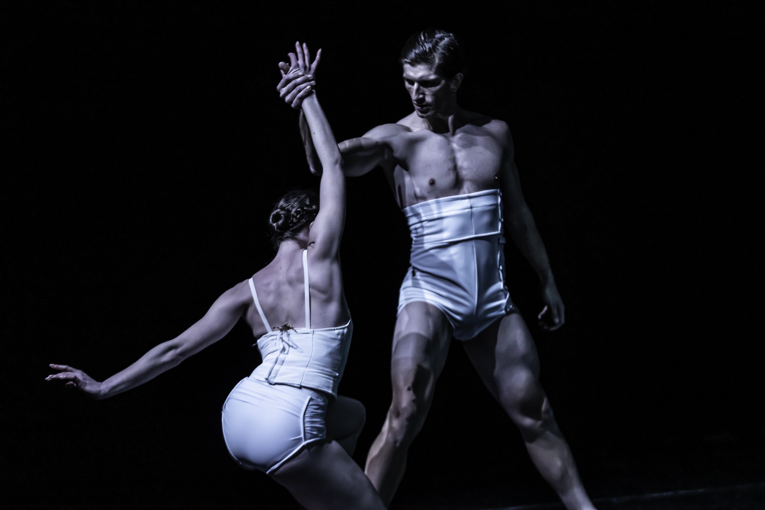 Two ballet dancers, a man and a woman, perform on stage wearing white costumes. The woman is kneeling and reaching up, while the man stands and holds her arm in a dance pose.