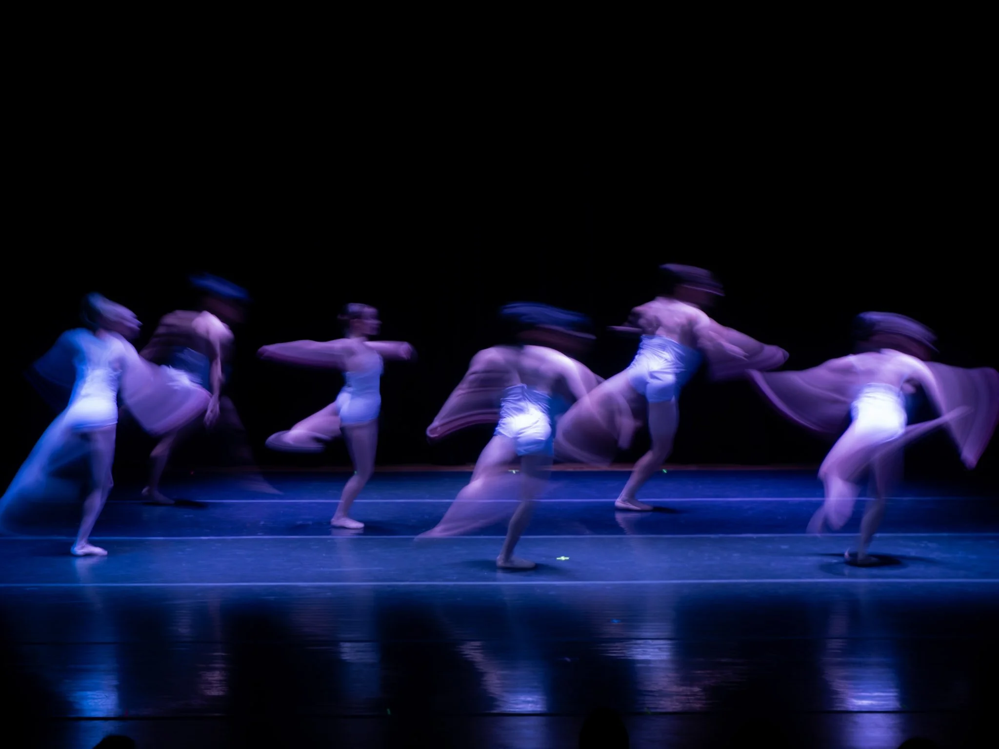A group of ballet dancers performing on stage with blue lighting, creating a blurred motion effect.