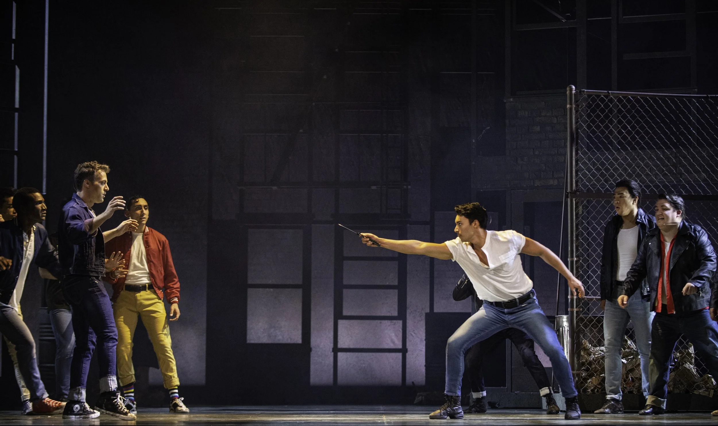 A scene from a theatrical performance shows a man in a white shirt and blue jeans pretending to sword fight with a man in a track jacket, surrounded by a group of people on stage. The stage has a dark industrial background and a chain-link fence.