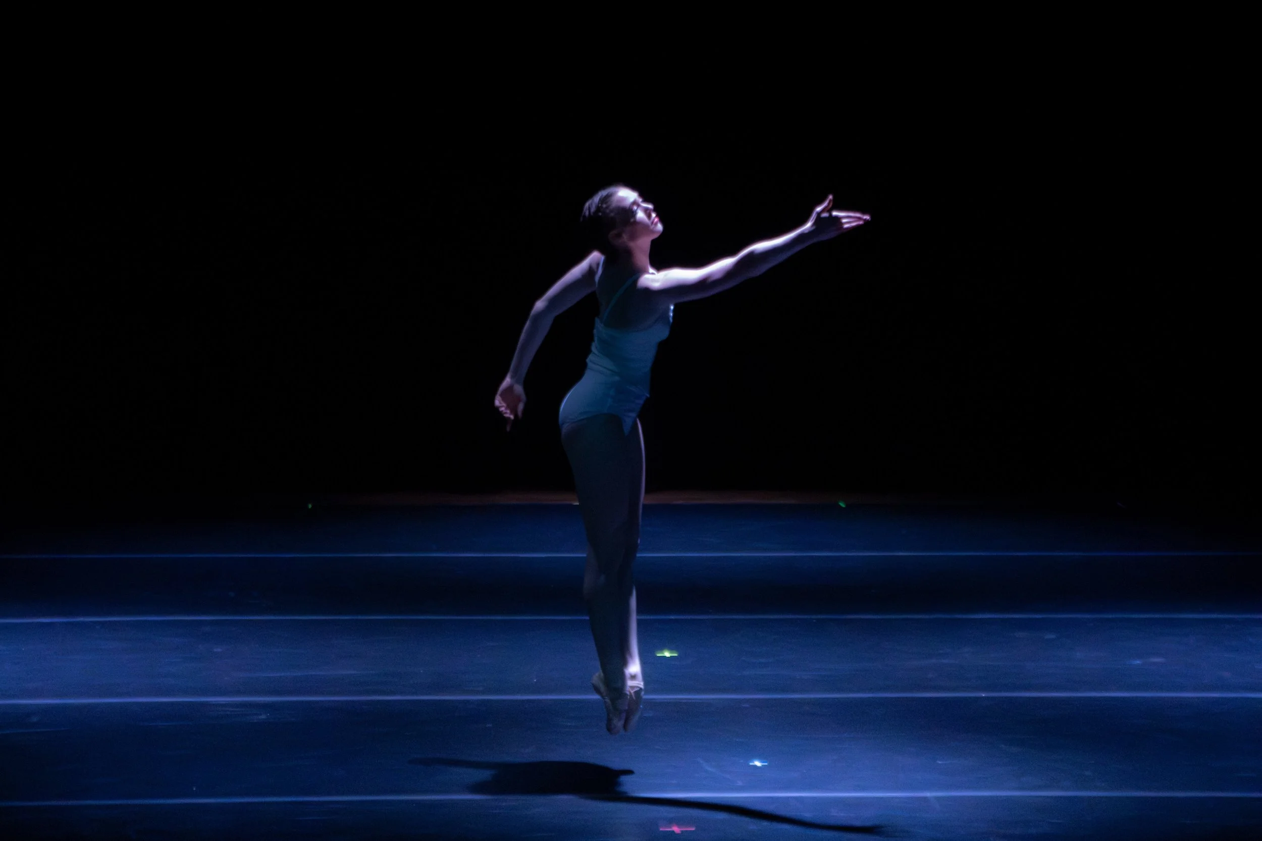 A female ballet dancer performing on a dark stage, illuminated by a spotlight, wearing a light-colored leotard and tights, mid-dance with an extended arm and raised on her toes.