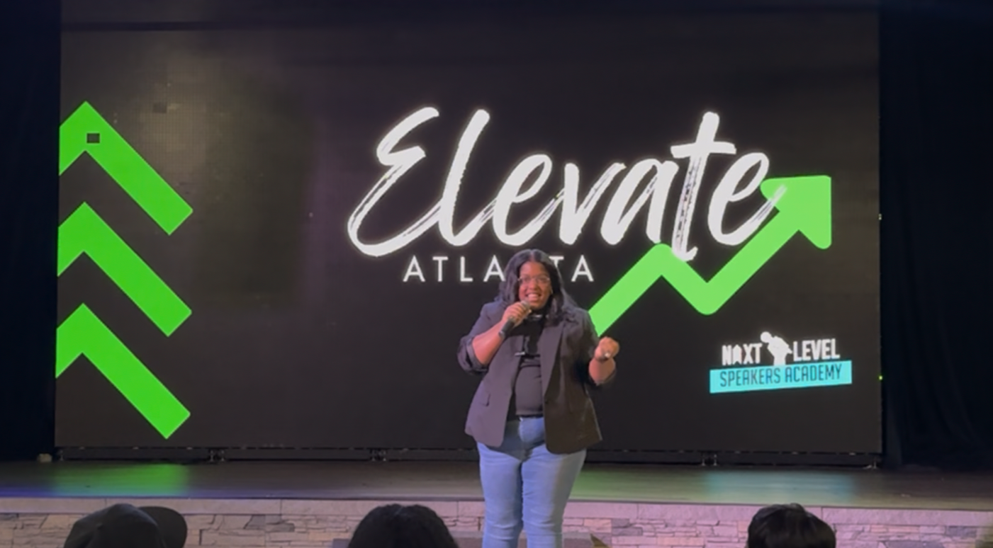 Woman speaking at a conference or event with a large screen behind her displaying the words 'Elevate Atlanta' and an upward green arrow.