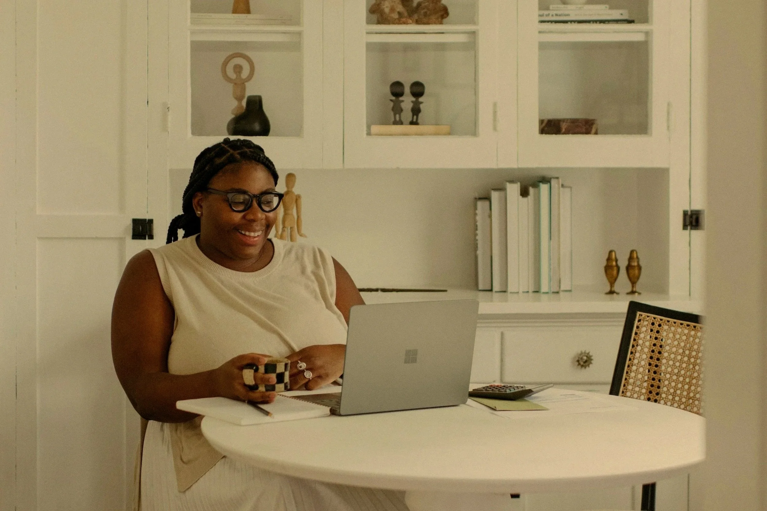 A woman with braided hair, glasses, and a sleeveless top smiling and looking at her laptop at a white table in a bright room with white cabinetry and decorative items on the shelves.