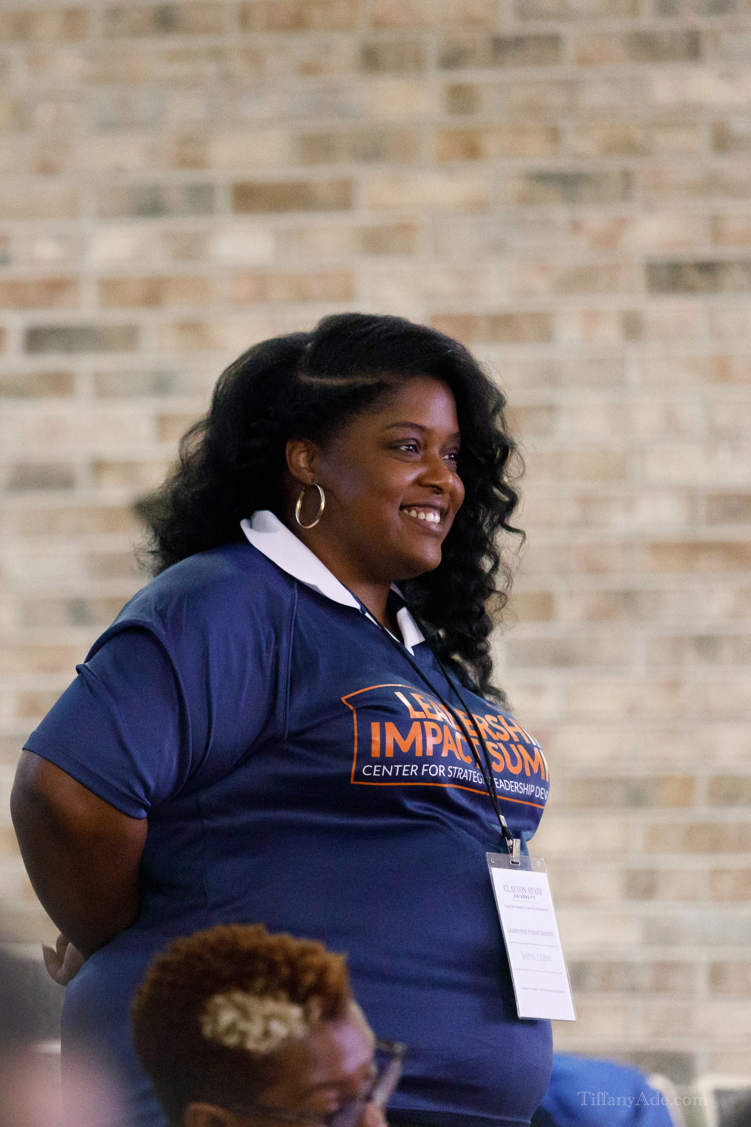 A smiling woman with dark, curly hair wearing a navy blue T-shirt with orange and white text, standing in front of a brick wall during an event.