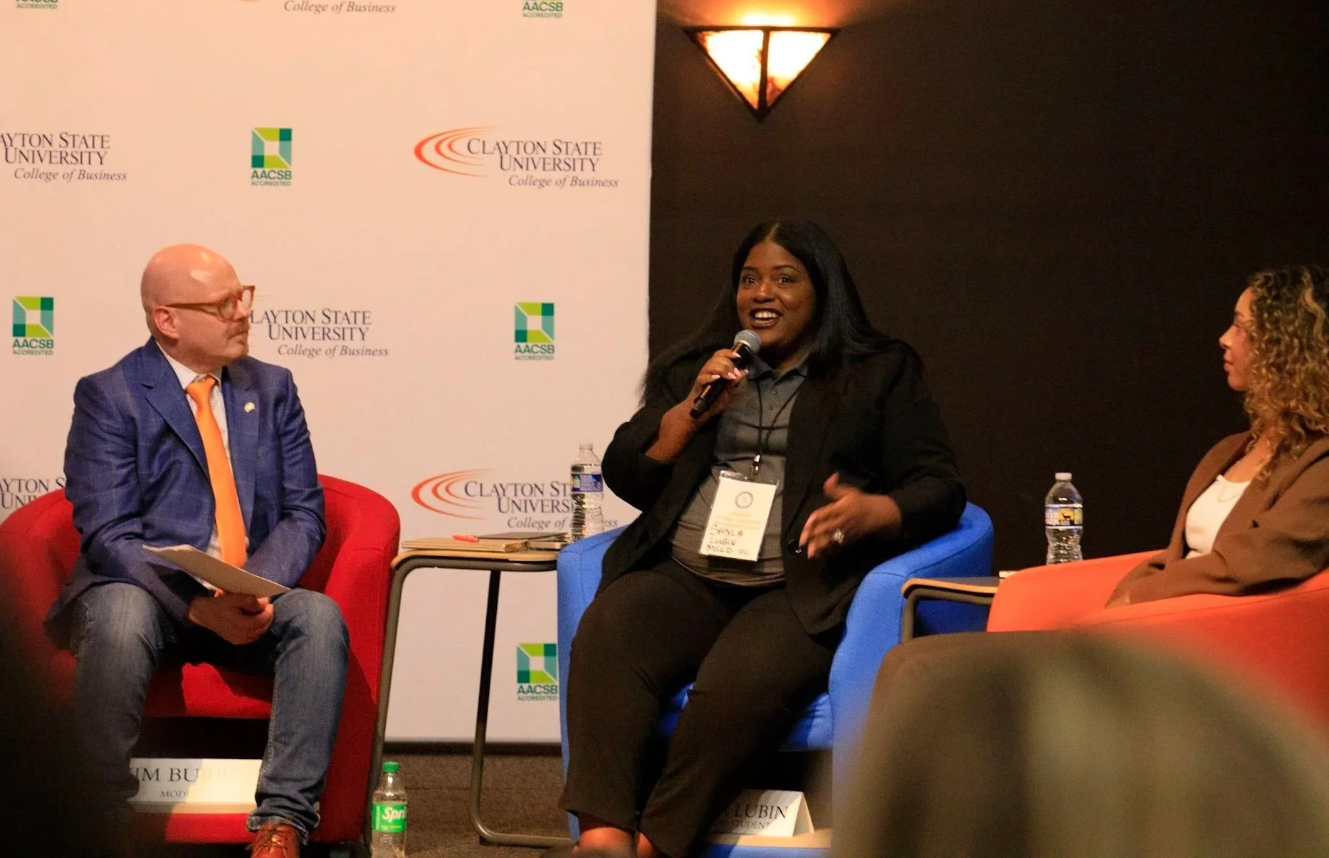 Three people seated on stage at a panel discussion with a white backdrop displaying logos for Clayton State University College of Business and AACSB. The woman in the middle is speaking into a microphone, with a water bottle on her side. The man on the left is dressed in a blue suit with an orange tie, and the woman on the right is wearing glasses and a brown blazer. All are seated on colorful chairs.