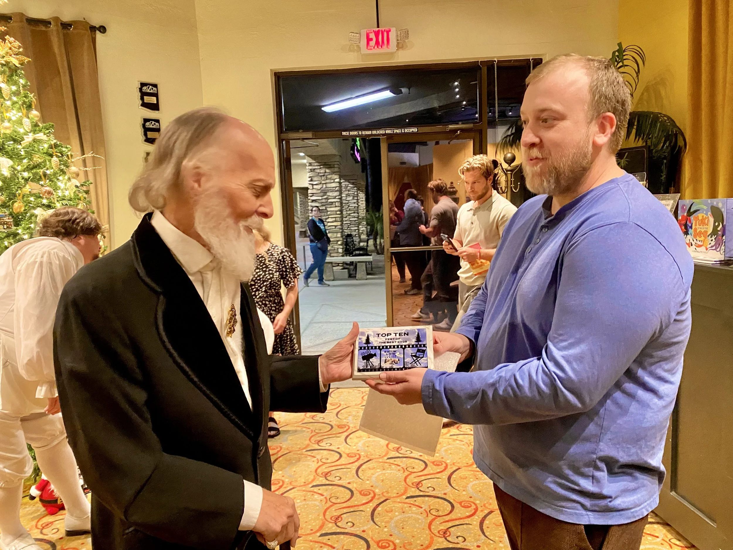 A man in a black jacket and white shirt is receiving a small box labeled 'Top Ten' from another man in a blue shirt at an indoor holiday event. A decorated Christmas tree and other people are visible in the background.
