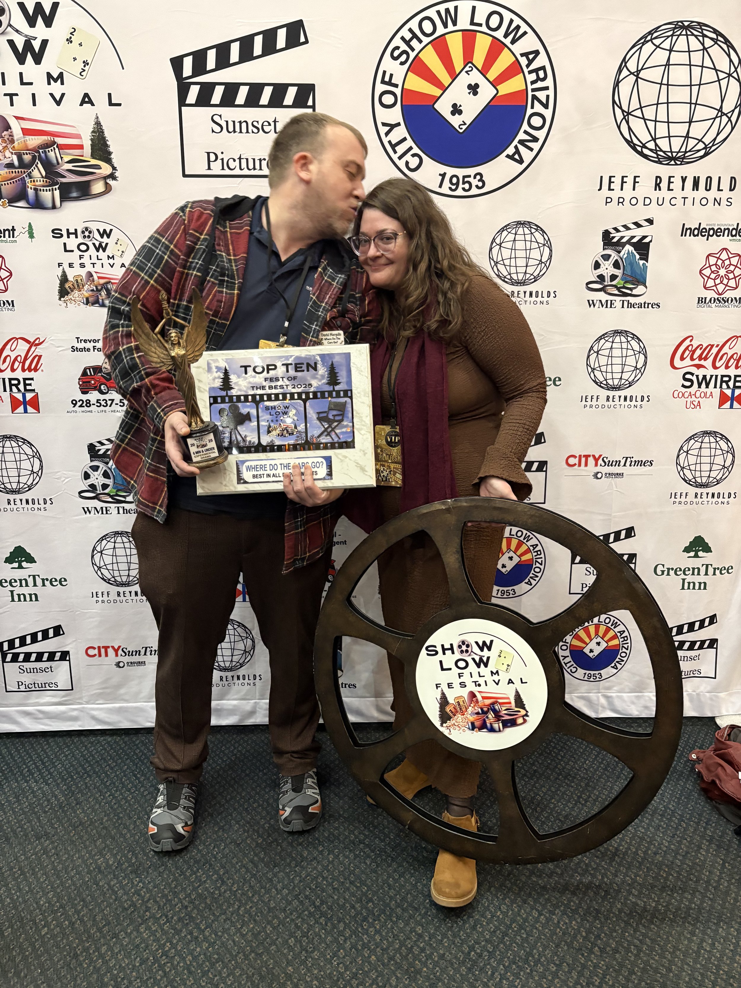 Two people stand in front of a backdrop with logos of the Show Low Film Festival and other sponsors, one holding a large trophy shaped like a film reel, and a box with the festival's logo. One person is kissing the other on the forehead.