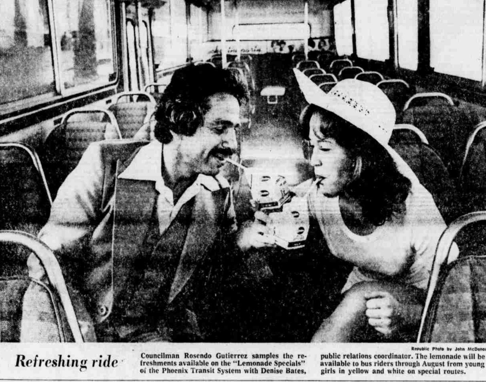 A black and white photograph of a man and woman sitting inside a bus, enjoying drinks through straws. The man is wearing a suit with a vest and has dark, curly hair. The woman is wearing a wide-brimmed hat and a light-colored blouse. Empty bus seats are visible in the background.