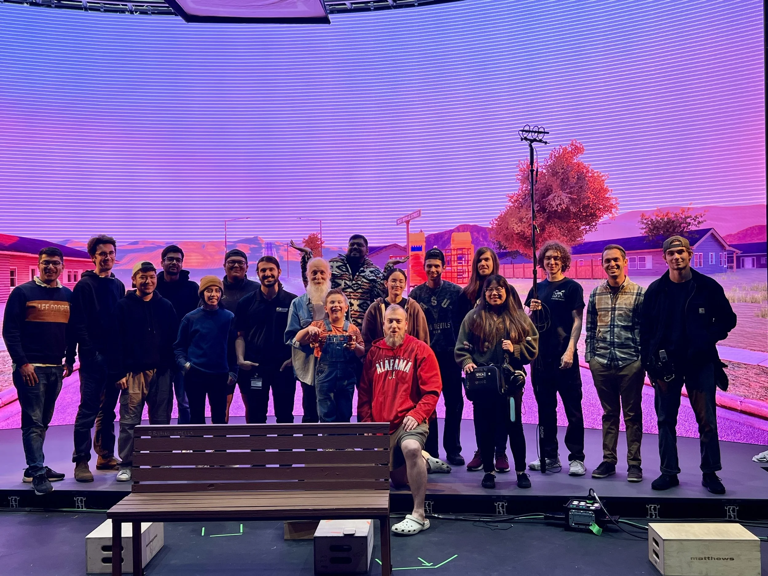Group of people, including children and adults, posing on a stage in front of a vibrant, animated background of a small town scene with houses, trees, and mountains during a presentation or event.