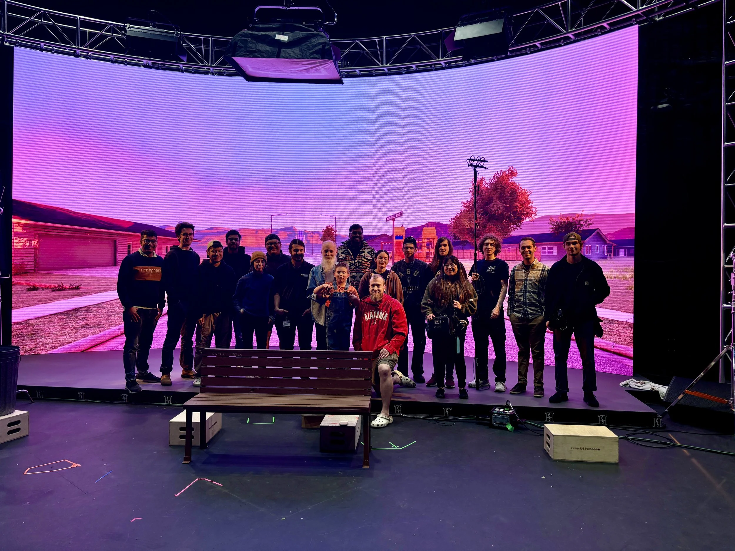 A group of people standing on a stage with a large digital screen behind them displaying a scenic landscape with purple and pink hues. The group includes men and women of various ages, some holding cameras or equipment, and one person kneeling in front wearing a red jacket. There is a bench and some equipment on the stage floor.
