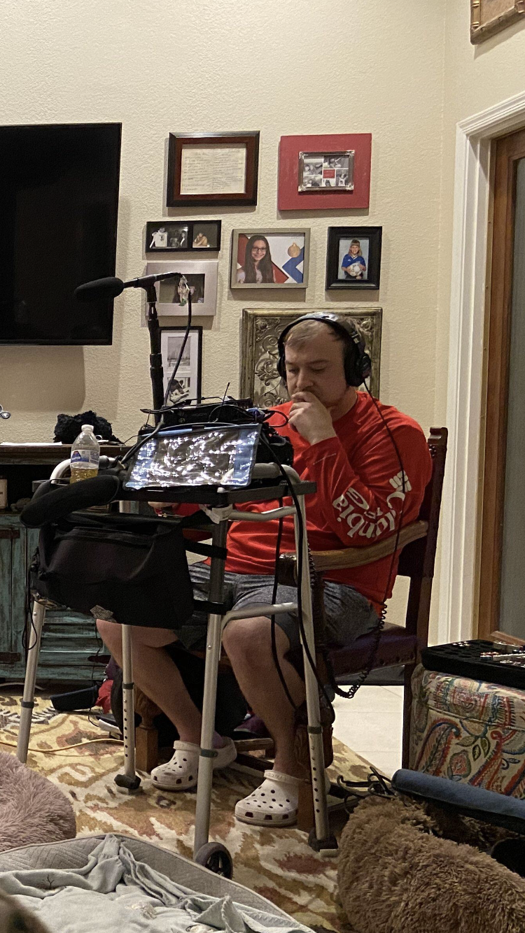A man sitting in a chair with a walker in a living room, wearing headphones, a red shirt, and Crocs, with a microphone setup in front of him and various electronic devices and a water bottle nearby. There are framed photos and decorations on the wall behind him.