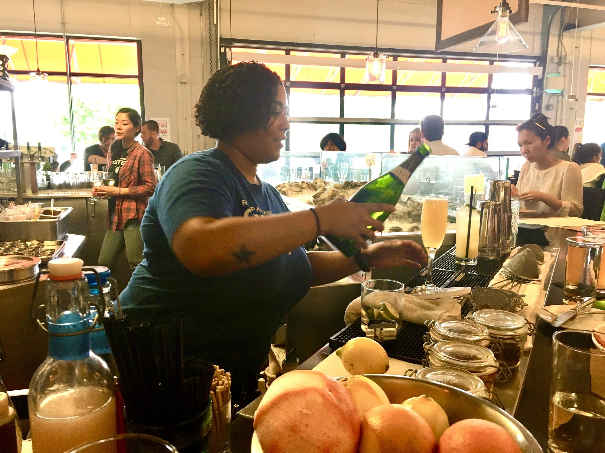 A bartender pours a drink behind a bar counter in a busy cafe or bar. People are seen sitting and standing, enjoying drinks in the background, with large windows letting in natural light.