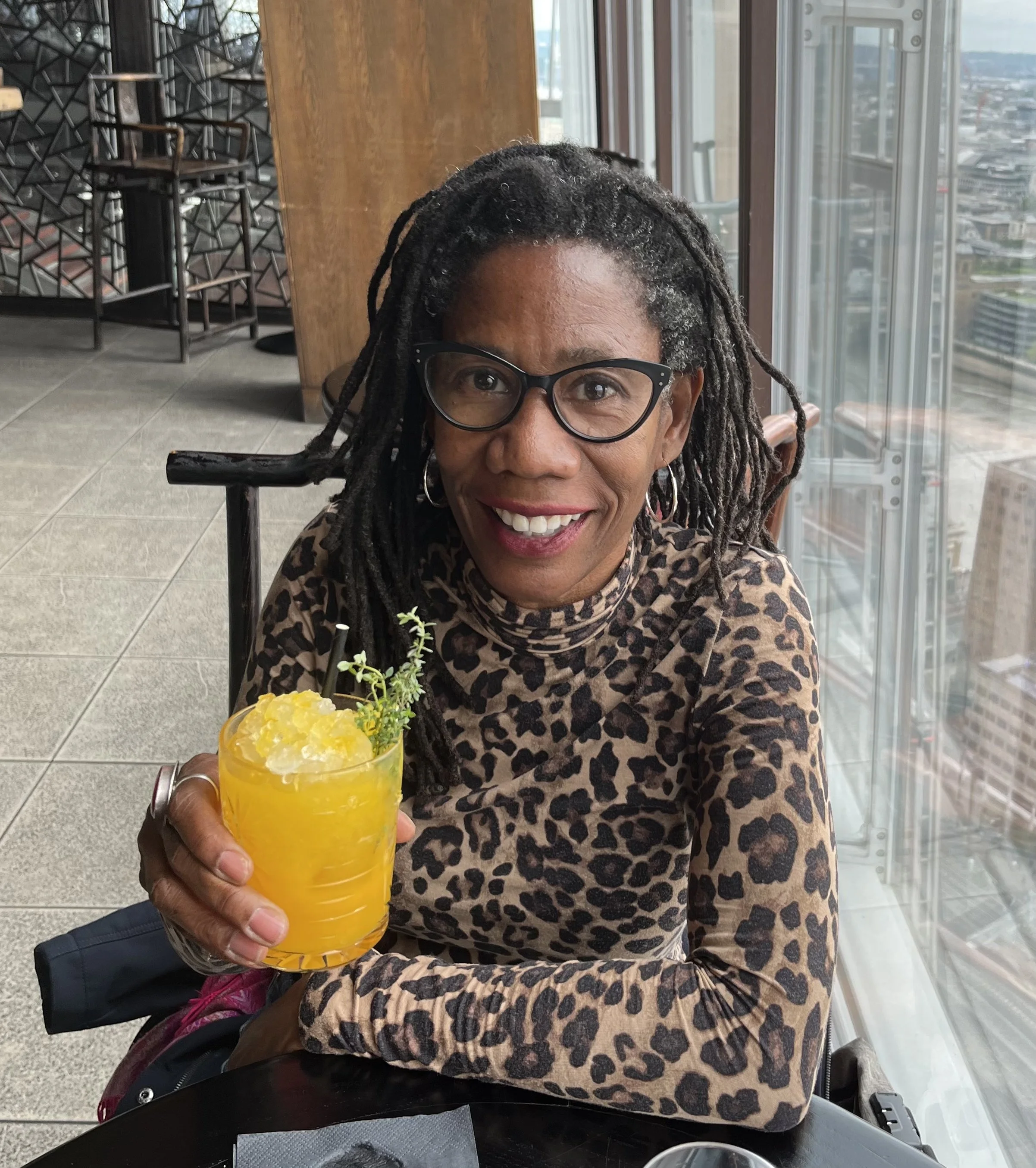 A smiling woman with dreadlocks, glasses, and hoop earrings, wearing a leopard print top, holding a yellow cocktail with crushed ice and garnished with a sprig of greenery, sitting at a table in a tall building restaurant with city view.