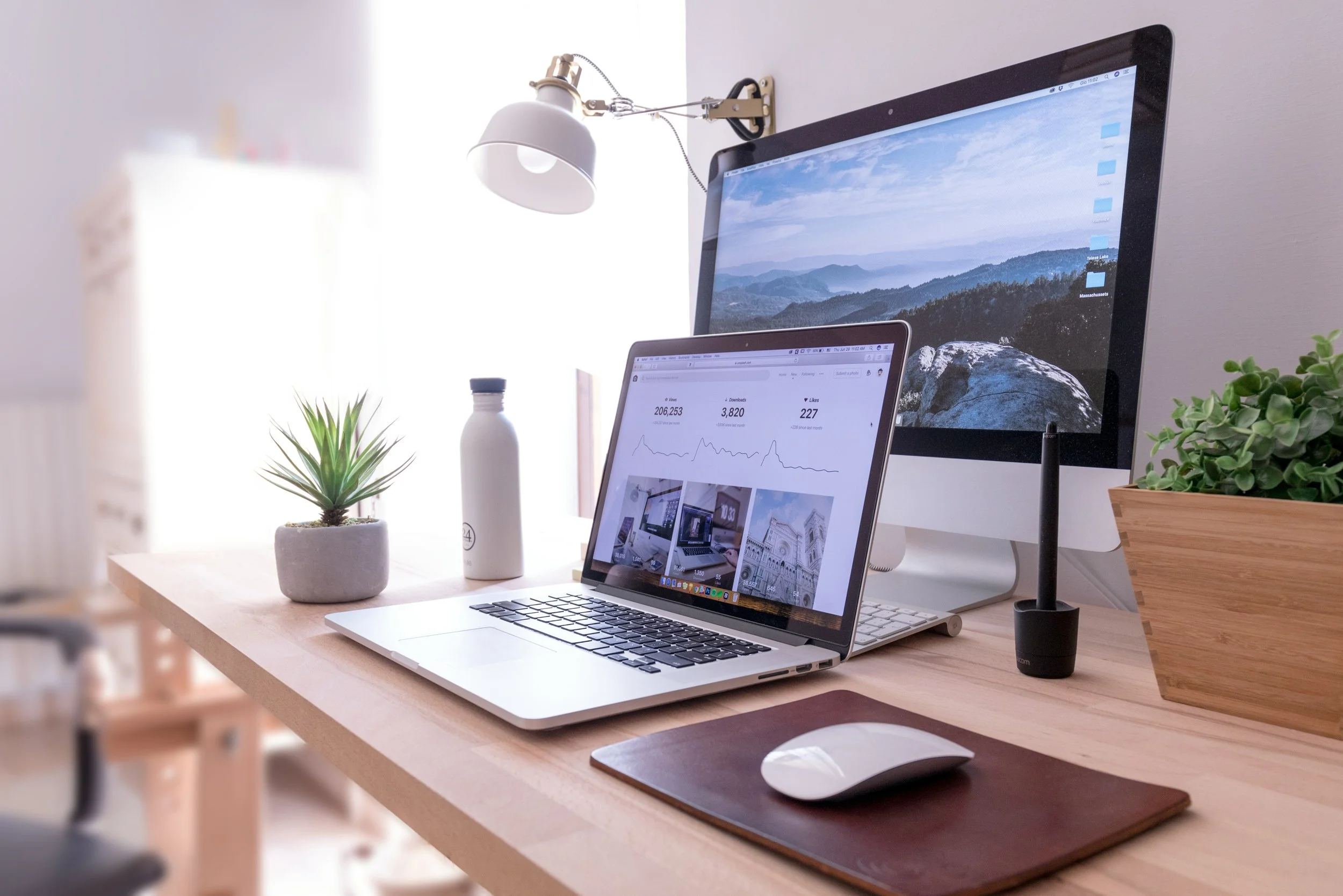 A clean workspace with a laptop, desktop monitor, mouse on a wooden desk, decorated with potted plants, a water bottle, and a desk lamp, in a bright room.