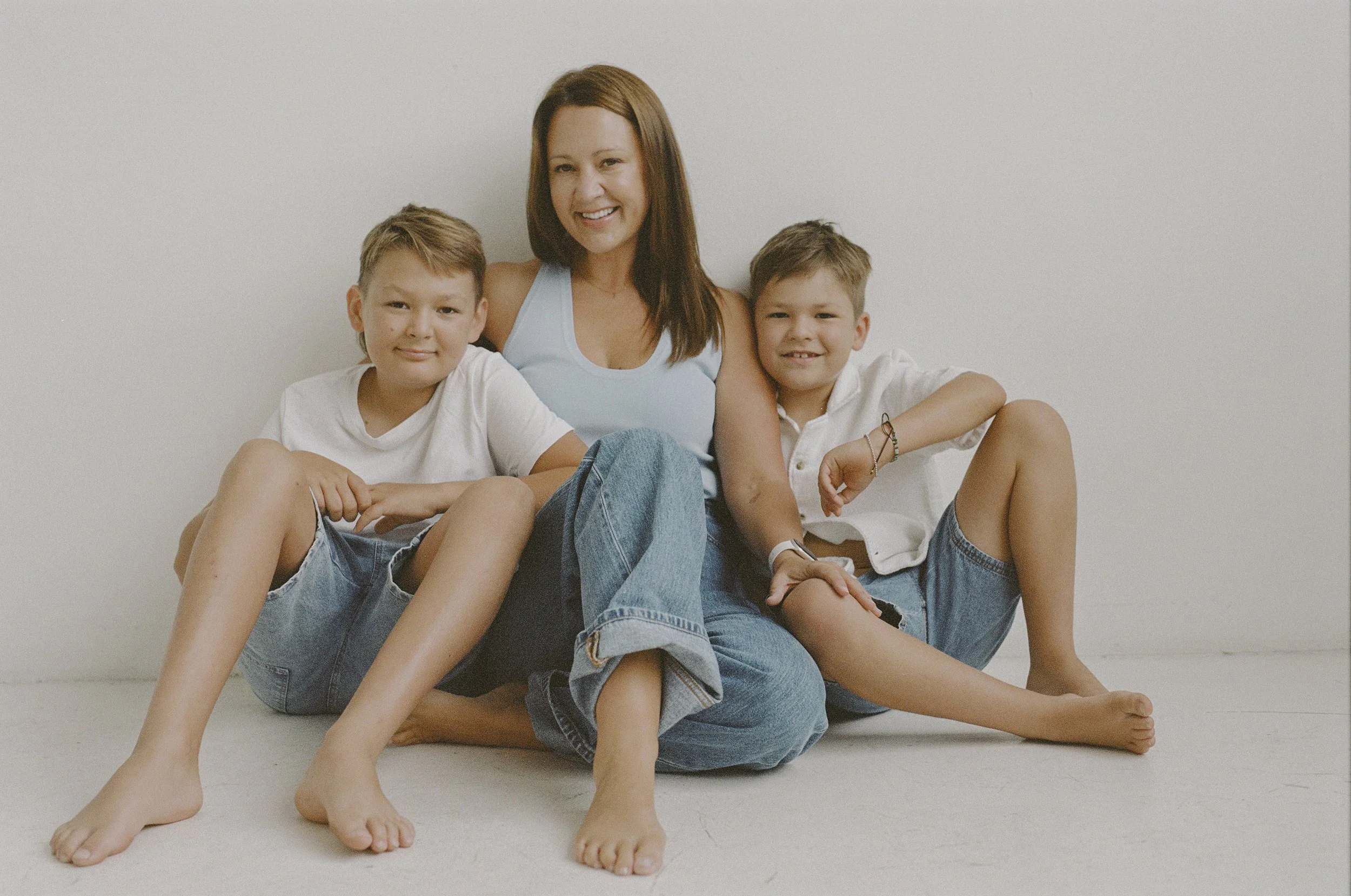 A woman sitting on the floor with two boys, all smiling. The woman is wearing a light blue tank top and jeans. The boys are dressed in white shirts and denim shorts, sitting close to the woman against a plain white background.