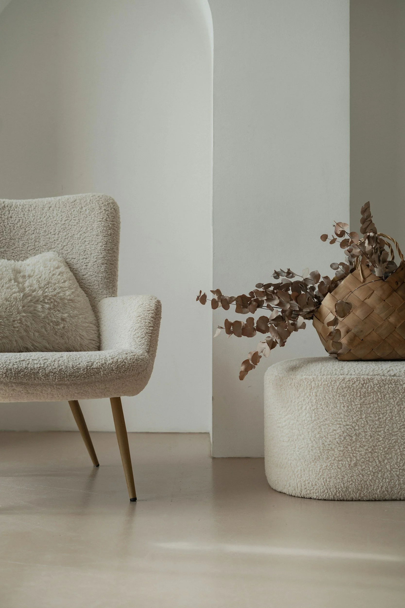 A beige armchair with wooden legs and textured upholstery, with a fluffy pillow on it, and a woven basket with dried eucalyptus branches, set against a minimalist white and gray wall.