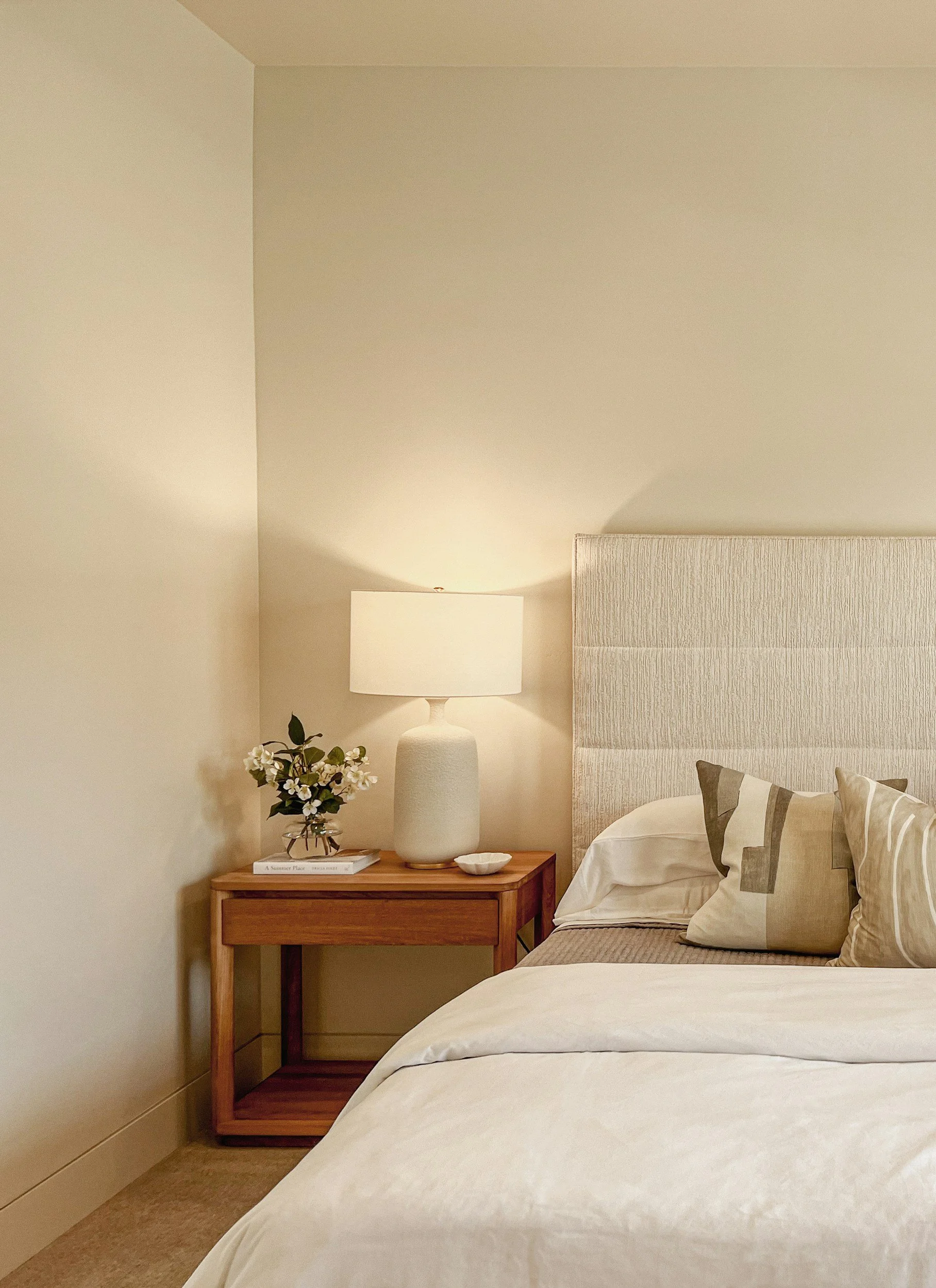 A bedroom with a bed, pillows, a bedside table with a lamp, a flower vase, and a small dish, with beige walls and a subtly textured headboard.