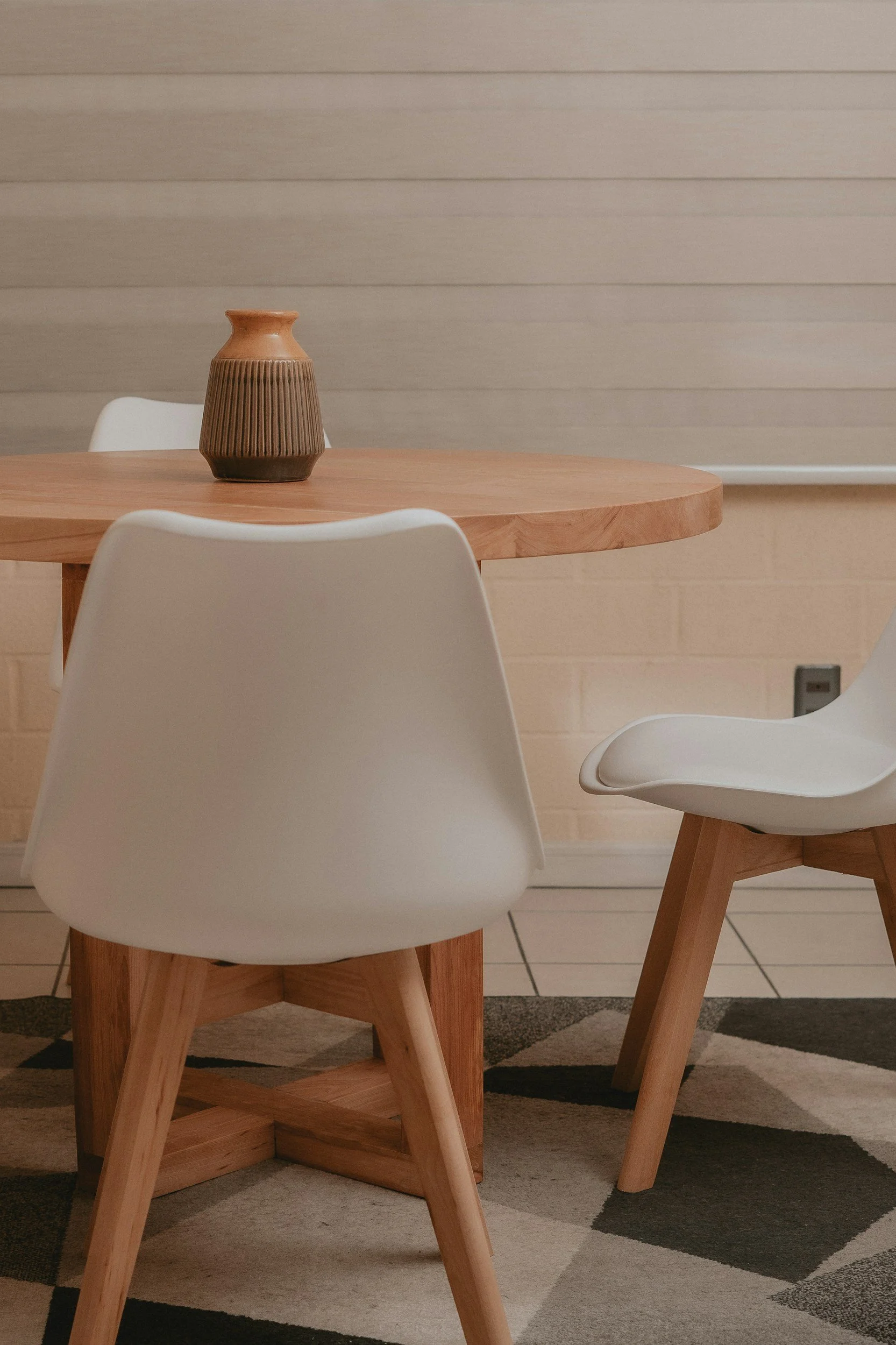 Modern dining area with a round wooden table, white chairs with wooden legs, a textured ceramic vase on the table, a light-colored wall with horizontal paneling, and a patterned rug on the floor.