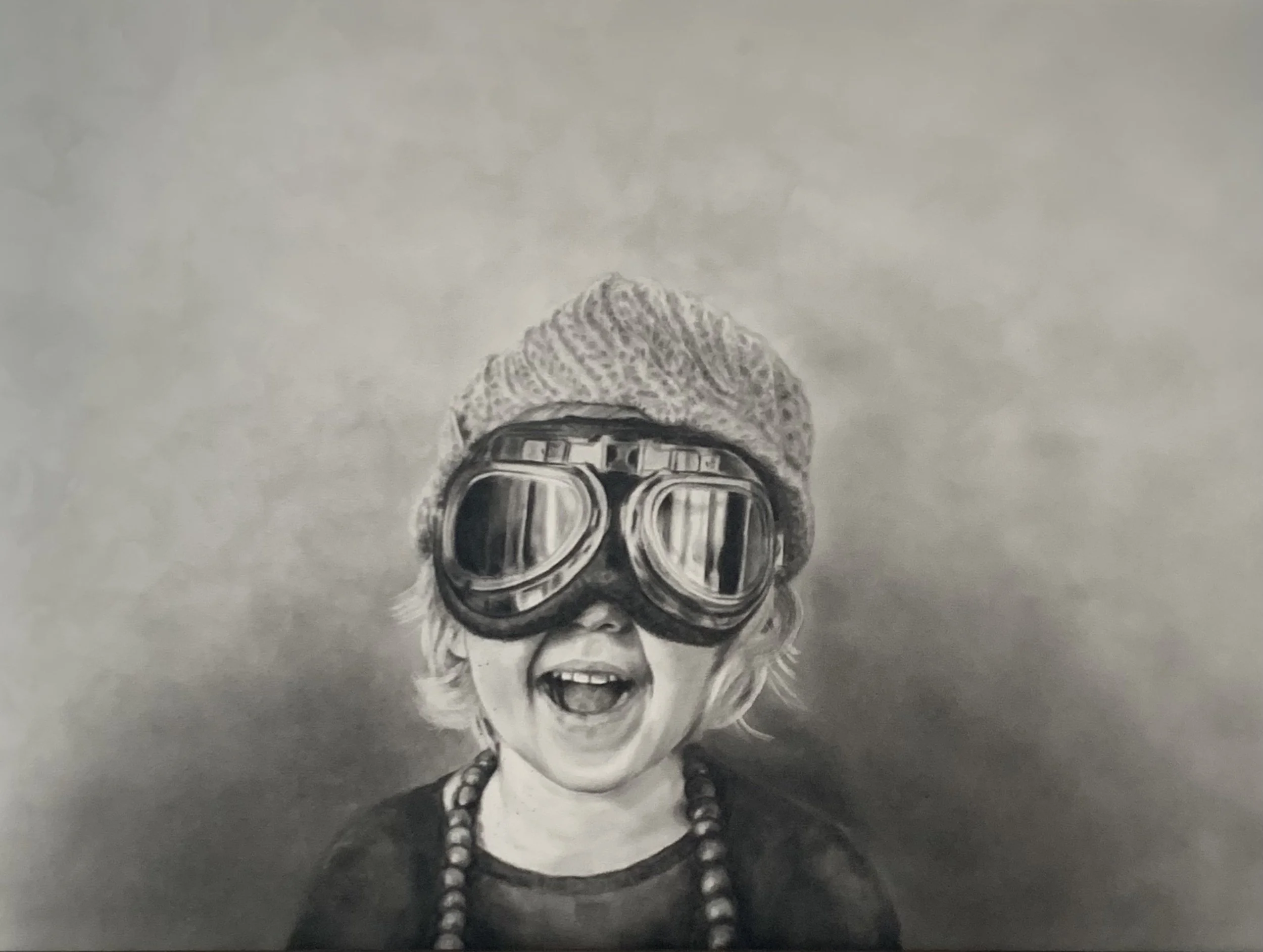 Black and white lifelike charcoal drawing of a child wearing large goggles, a knitted hat, and a beaded necklace, with a joyful expression.