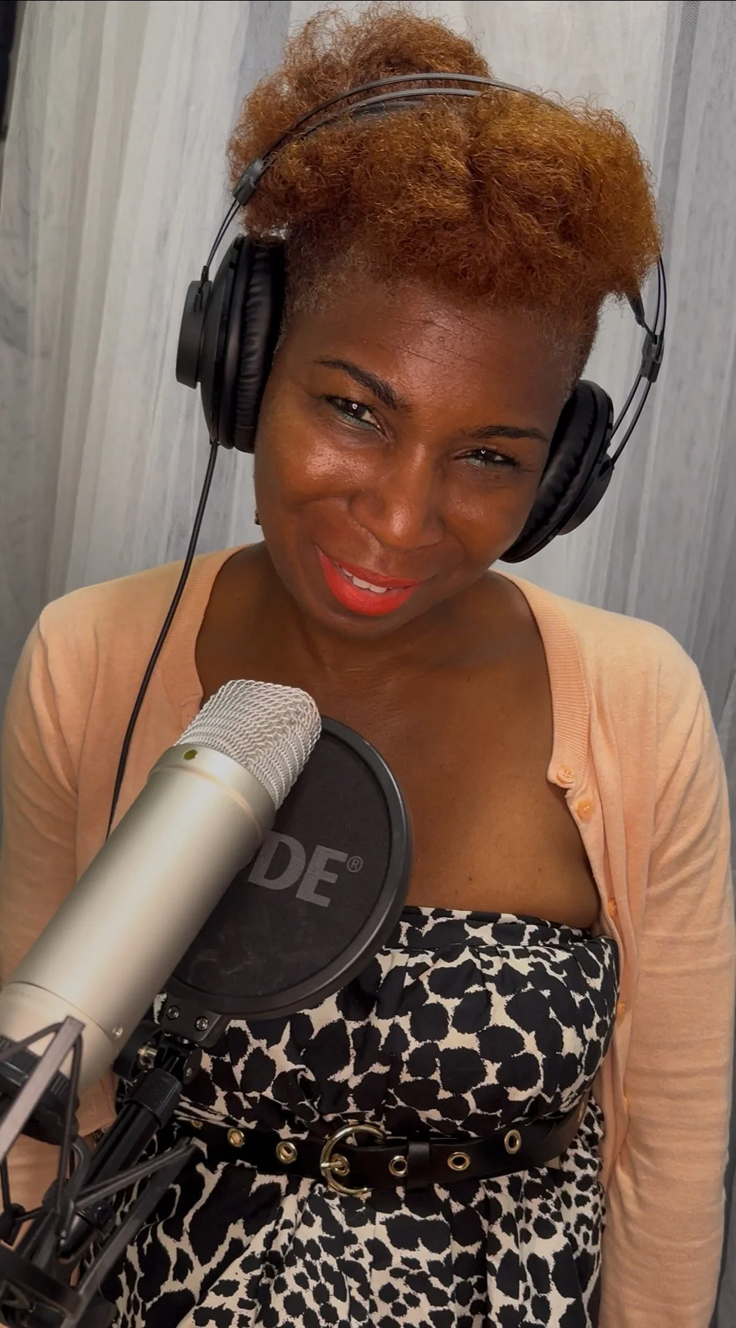 Karen Joseph the voice of health and wellness, with curly orange hair wearing headphones, standing in front of a microphone in a recording studio. Karen Joseph the voice of health and wellness, voice-acting, voice actor.