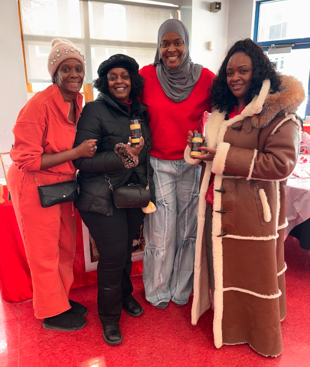 Four women posing indoors, smiling, with three of them holding candles. They are dressed warmly, with one wearing a pink hat, another in a black jacket, the third in a red sweater, and the fourth in a brown shearling coat. Behind them are large windows and a table with a pink tablecloth.