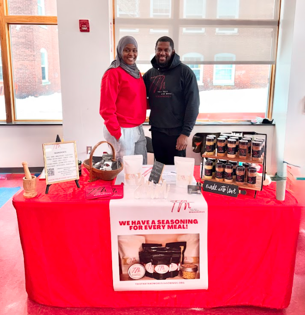 Two people standing behind a booth table with a red tablecloth, displaying jars of seasoning and spices, inside a well-lit room with large windows and a brick building outside.