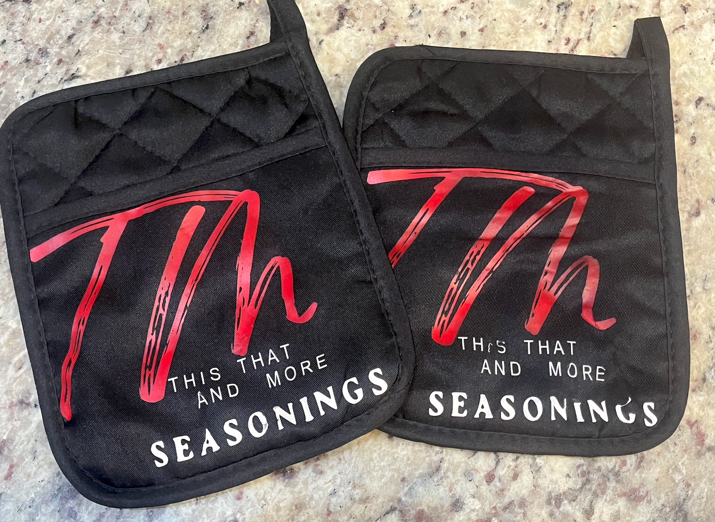 Two black cloth oven mitts with quilted pattern and red and white print, hanging on a speckled countertop.