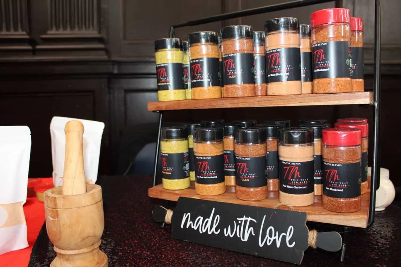 Display of various spice jars on a two-tier wooden rack with a sign reading 'made with love' in front, and a wooden mortar and pestle on the table.