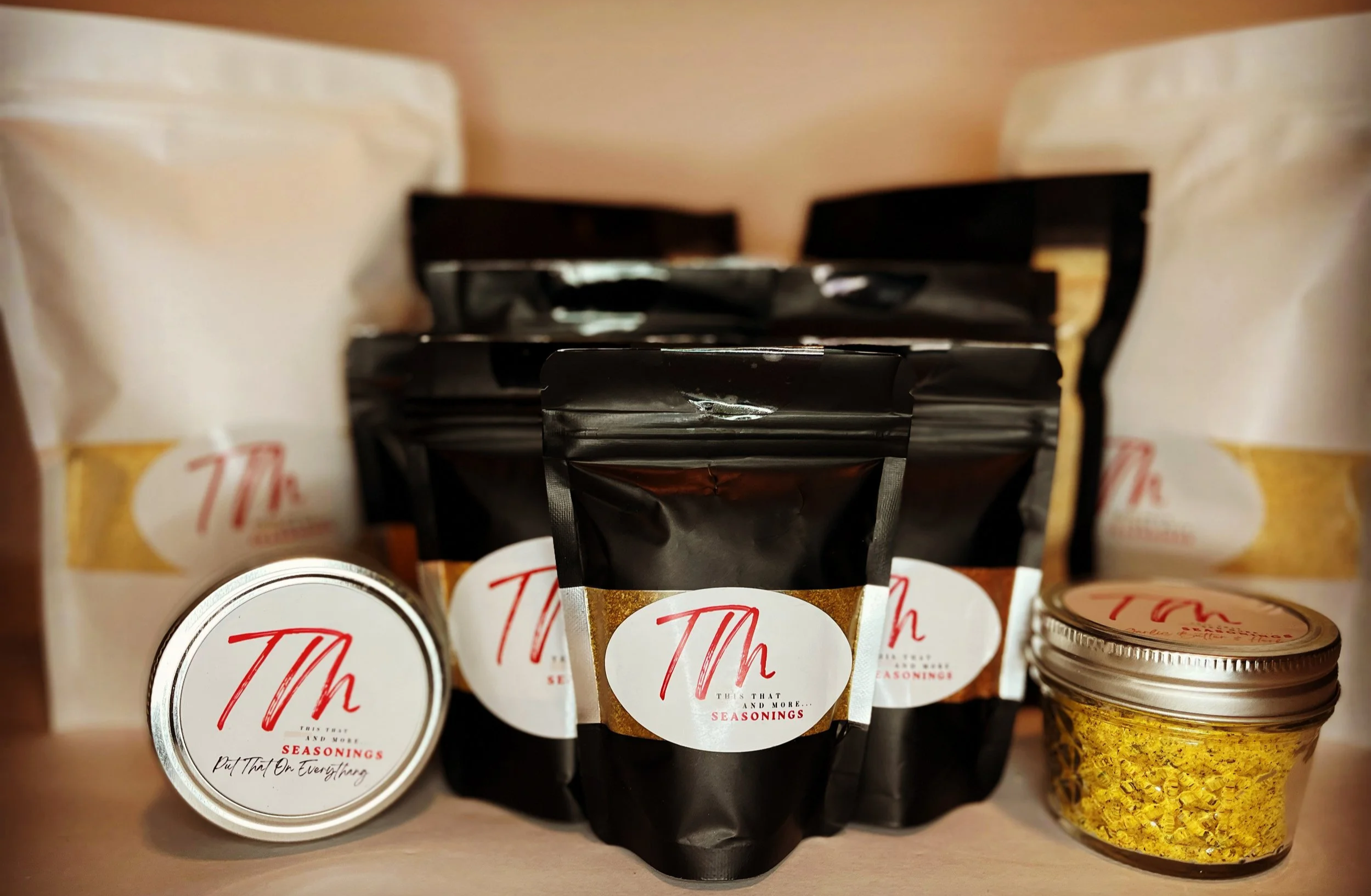 Various seasonings in black resealable pouches with gold and white labels, a small jar with yellow seasoning, and two white bags with similar labels, all labeled "Th".