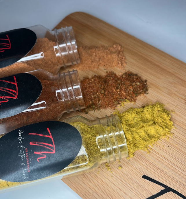 Three glass spice jars with black labels labeled 'V,' containing brown, red, and yellow spice powders spilled onto a wooden cutting board.