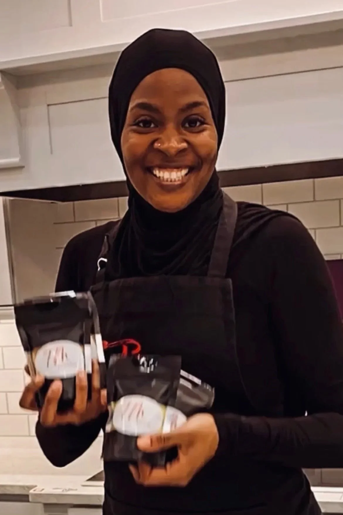 A smiling woman wearing a black hijab and apron, holding multiple packets of face masks in a kitchen setting.