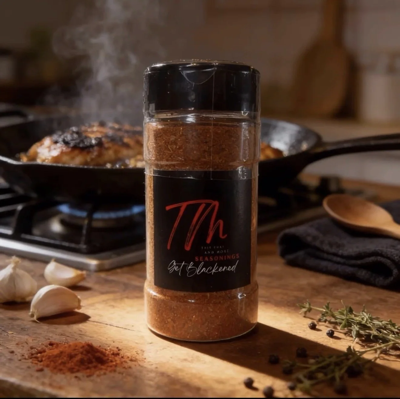A spice jar labeled "Th" with the tagline "Get Blackened" beside garlic cloves, black peppercorns, and thyme on a wooden kitchen countertop. A cast iron skillet with steaming food is on a stove in the background.