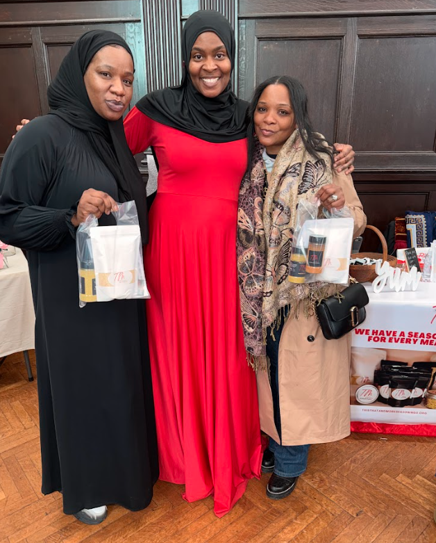 Three women standing together indoors, smiling, holding gift bags. One in a black dress and hijab, another in a red dress and hijab, the third in a beige coat with a colorful scarf. Background includes tables with prizes and a sign.